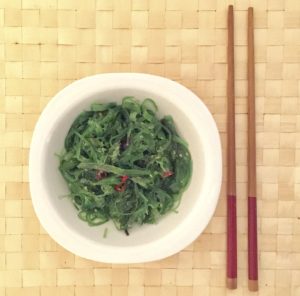 Ensalada de algas wakame con sésamo y chile. Se presenta en un bol blanco sobre yn mantillo vegetal trenzado y unos palillos orientales.