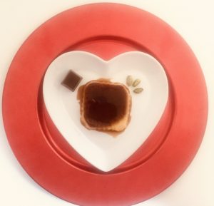 Tocino de cielo con chocolate y pistachos. Tiene forma cuadrada y está presentado sobre un plato en forma de corazón blanco y un sobre plato rojo. Se observa un bombón de chocolate y unos pistachos tostados pelados. Menú de San Valentín rápido y resultón