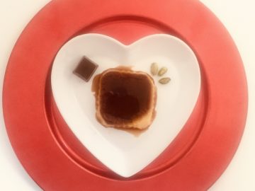 Tocino de cielo con chocolate y pistachos. Tiene forma cuadrada y está presentado sobre un plato en forma de corazón blanco y un sobre plato rojo. Se observa un bombón de chocolate y unos pistachos tostados pelados.