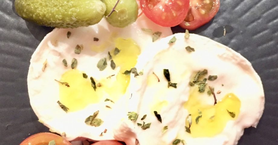 Tapa de mozzarella con cherry y pepinillos. Sobre un plato negro con aceite y orégano.